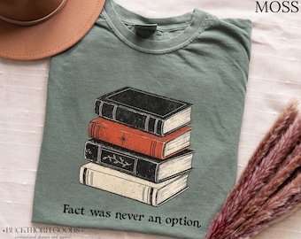 Könnte beinhalten: Ein moosgrünes T-Shirt mit einer Grafik eines Stapels aus vier Büchern in den Farben Schwarz, Orange und Creme. Das orangefarbene Buch hat ein Sternsymbol und das schwarze Buch darunter ein Blattmotiv. Der Text "Fact was never an option" ist darunter aufgedruckt.