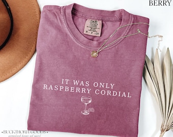 Könnte beinhalten: Ein beerenfarbenes T-Shirt mit Rundhalsausschnitt und dem weißen Schriftzug IT WAS ONLY RASPBERRY CORDIAL über einer kleinen Grafik eines Stielglases. Das Shirt hat ein Comfort Colors Markenetikett im Kragen und eine weiche, verwaschene Textur.