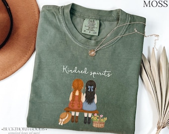 Könnte beinhalten: Ein moosgrünes T-Shirt mit einer Grafik von zwei Figuren auf einer Bank, eine mit roten Zöpfen und eine mit dunklem Haar. Der Text Kindred spirits steht darüber. Das T-Shirt hat ein Comfort Colors Etikett in der Größe S.
