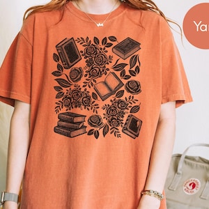 May include: A short-sleeve crew neck t-shirt in a solid burnt orange color. The front features a black graphic print consisting of several books and clusters of roses with leaves arranged in a scattered pattern across the center of the chest.