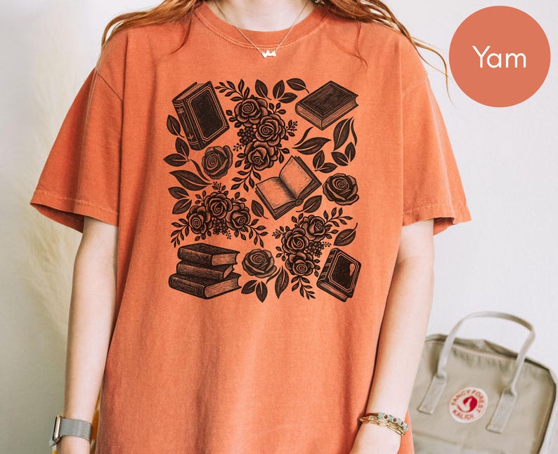 May include: A short-sleeve crew neck t-shirt in a solid burnt orange color. The front features a black graphic print consisting of several books and clusters of roses with leaves arranged in a scattered pattern across the center of the chest.