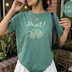 Puede incluir: Una camiseta de manga corta de color verde claro con un gr&aacute;fico de color crema de dos jarras de cerveza chocando entre s&iacute;, con el texto "Prost!" impreso encima. El dise&ntilde;o est&aacute; centrado en el pecho de la prenda, que tiene cuello redondo.