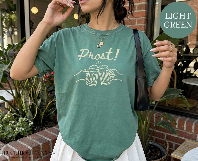 Puede incluir: Una camiseta de manga corta de color verde claro con un gr&aacute;fico de color crema de dos jarras de cerveza chocando entre s&iacute;, con el texto "Prost!" impreso encima. El dise&ntilde;o est&aacute; centrado en el pecho de la prenda, que tiene cuello redondo.