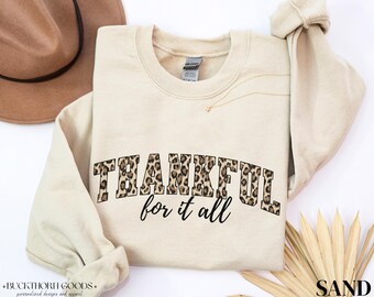 Könnte beinhalten: Ein sandfarbenes Sweatshirt mit Rundhalsausschnitt, das das Wort THANKFUL in einem braun-schwarzen Leopardenmuster zeigt, mit den Worten for it all in einer schwarzen Schreibschrift unter dem Haupttext. Das Kleidungsstück hat lange Ärmel.