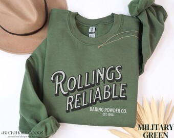 Könnte beinhalten: Ein militärgrüner Rundhals-Pullover mit dem schwarz-weißen Schriftzug Rollings Reliable Baking Powder Co. Est. 1908. Das Kleidungsstück hat lange Ärmel und einen gerippten Kragen, wobei ein beigefarbener Hut und getrocknete Palmblätter am Rand zu sehen sind.
