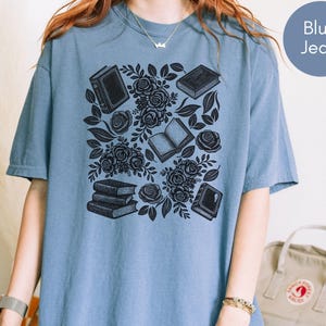May include: A blue jean colored t-shirt featuring a black graphic print of several books and blooming roses arranged in a cluster. The design includes both open and closed books interspersed with detailed floral elements and leaves across the center.