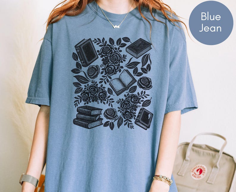 May include: A blue jean colored t-shirt featuring a black graphic print of several books and blooming roses arranged in a cluster. The design includes both open and closed books interspersed with detailed floral elements and leaves across the center.
