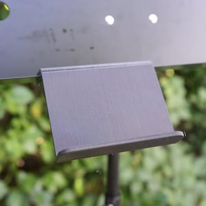Könnte beinhalten: Ein schwarzer Notenständer aus Metall mit einer rechteckigen Notenablage. Der Ständer befindet sich vor einem verschwommenen grünen Hintergrund aus Blättern. Die Notenablage ist leicht nach oben abgewinkelt.