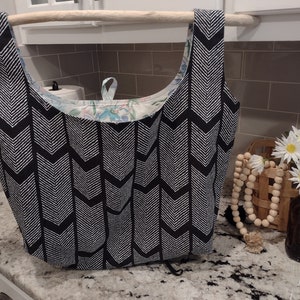 May include: A black and white chevron patterned reusable shopping bag with a floral patterned lining. The bag is hanging from a wooden dowel.