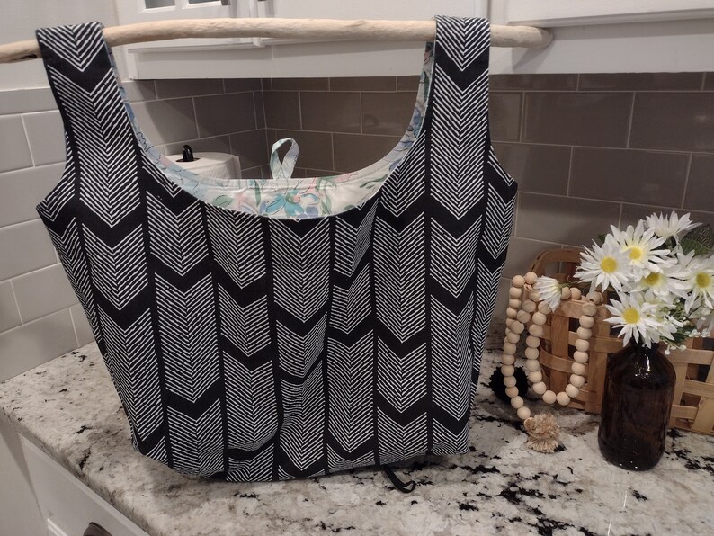 May include: A black and white chevron patterned reusable shopping bag with a floral patterned lining. The bag is hanging from a wooden dowel.