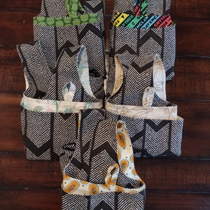May include: Six reusable shopping bags folded and stacked on a wooden surface. The bags are made of black and white fabric with a chevron pattern. The top bag is green with white polka dots. The second bag has a colourful pattern with the text "The Other Side of England" and "Ravenclaw Gryffindor".