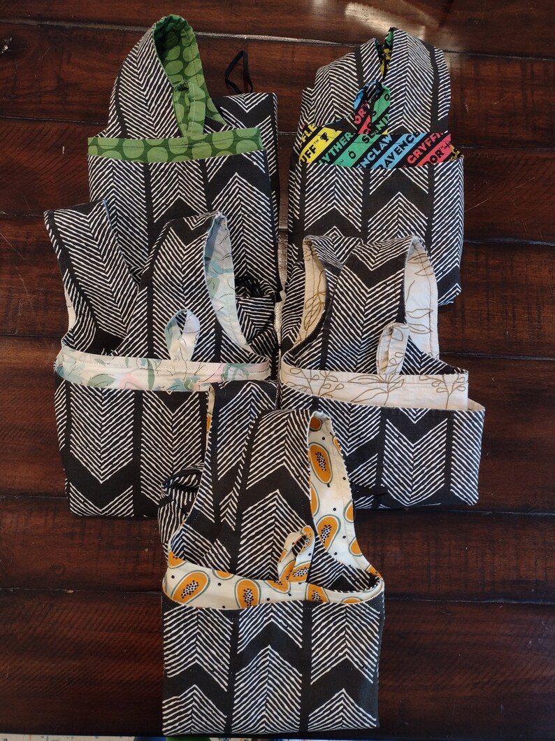 May include: Six reusable shopping bags folded and stacked on a wooden surface. The bags are made of black and white fabric with a chevron pattern. The top bag is green with white polka dots. The second bag has a colourful pattern with the text "The Other Side of England" and "Ravenclaw Gryffindor".