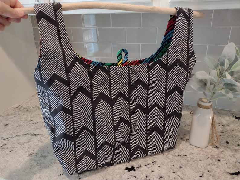 May include: A black and white chevron patterned reusable shopping bag with a colourful fabric lining. The bag is hanging from a wooden dowel.