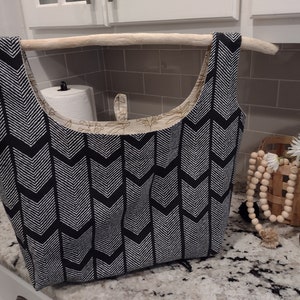 May include: A black and white chevron patterned tote bag with a wooden dowel rod handle. The bag is hanging on a shelf in a kitchen.