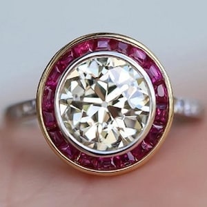 Peut inclure: Une bague de fiançailles en diamant rond avec un halo de rubis sertis dans de l'or jaune. La bague a une bande en or blanc avec de petits diamants.