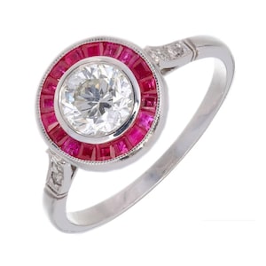 Peut inclure: Une bague de fiançailles en platine avec un diamant central rond brillant entouré d'un halo de rubis rouges. La bague a une bande simple avec de petits diamants sur les côtés.