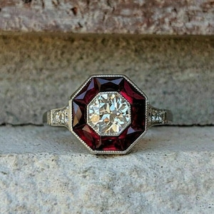 Puede incluir: Un anillo de estilo vintage con una gran piedra central clara y facetada, rodeada por un marco octogonal rojo oscuro. La banda plateada tiene pequeñas piedras de acento a cada lado de la piedra central.