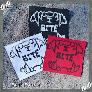 以下が含まれることがあります： 黒、白、赤の3つの正方形のパッチ。それぞれに様式化された顎のデザインと、黒で「I BITE」の文字が描かれています。パッチは、テクスチャ加工された灰色の表面に配置されています。「BITE PATCH」の文字が下にあります。