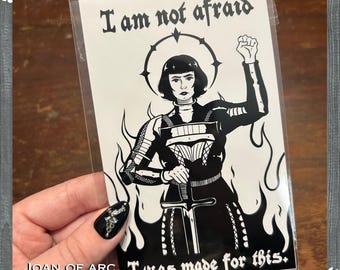 May include: A black and white print of Joan of Arc, with the text "I am not afraid" and "I was made for this." The image shows Joan of Arc holding a sword, with flames around her, and a halo. The print is labelled "JOAN OF ARC PRINT."
