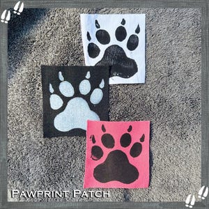 以下が含まれることがあります： 足跡のデザインが施された正方形のパッチが3つ。1つは黒い足跡の白いパッチ、もう1つは白い足跡の黒いパッチ、3つ目は黒い足跡のピンクのパッチです。「PAWPRINT PATCH」の文字が下にあります。
