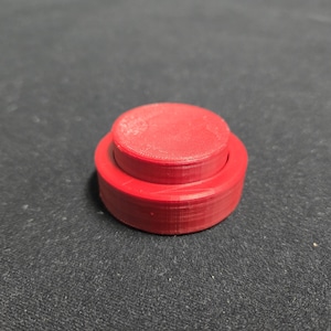 May include: A small, red, cylindrical object with a stepped design. The top portion is a smaller, circular disc, and the base is a wider, circular ring. The object is made of a solid material and has a matte finish.