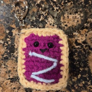 May include: Crocheted keychain in the shape of a Pop-Tart. The Pop-Tart has a purple filling with a white zigzag design, black eyes, and a small smile. The edges are tan, and it has a silver keychain ring.