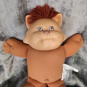 Peut inclure: Une poupée en peluche de chat marron avec une crinière en laine rouge et de grands yeux verts. La poupée a un nez et une bouche cousus et est allongée sur le dos, les bras tendus.