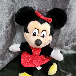 Puede incluir: Un juguete de peluche de Minnie Mouse con un vestido rojo de lunares blancos y un lazo rojo. Tiene orejas negras y una nariz negra. Sus ojos son grandes y marrones.
