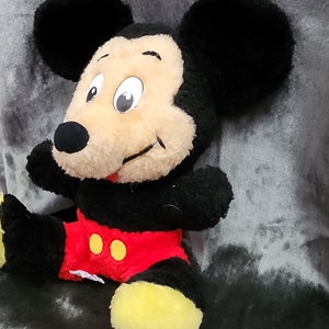 May include: A plush Mickey Mouse toy with black ears, a black body, red shorts with yellow buttons, and yellow shoes. The toy is sitting on a grey surface.