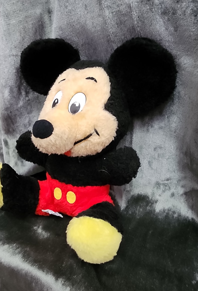 May include: A plush Mickey Mouse toy with black ears, a black body, red shorts with yellow buttons, and yellow shoes. The toy is sitting on a grey surface.