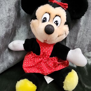 Puede incluir: Un juguete de peluche de Minnie Mouse con un vestido rojo con lunares blancos y un lazo rojo. El juguete tiene orejas negras, una nariz negra y pies amarillos.