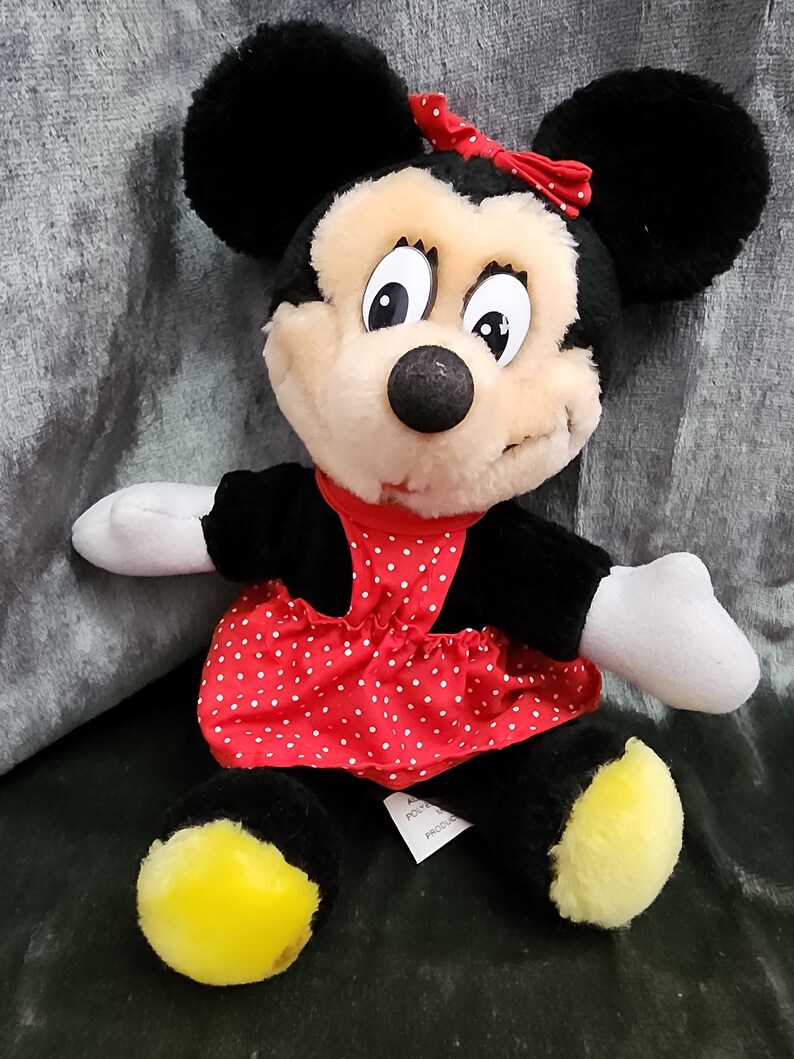 Puede incluir: Un juguete de peluche de Minnie Mouse con un vestido rojo con lunares blancos y un lazo rojo. El juguete tiene orejas negras, una nariz negra y pies amarillos.