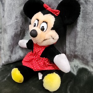 Puede incluir: Un juguete de peluche Minnie Mouse negro y tostado con un vestido rojo de lunares y un lazo rojo. El juguete tiene pies amarillos y est&aacute; sentado en una superficie gris.