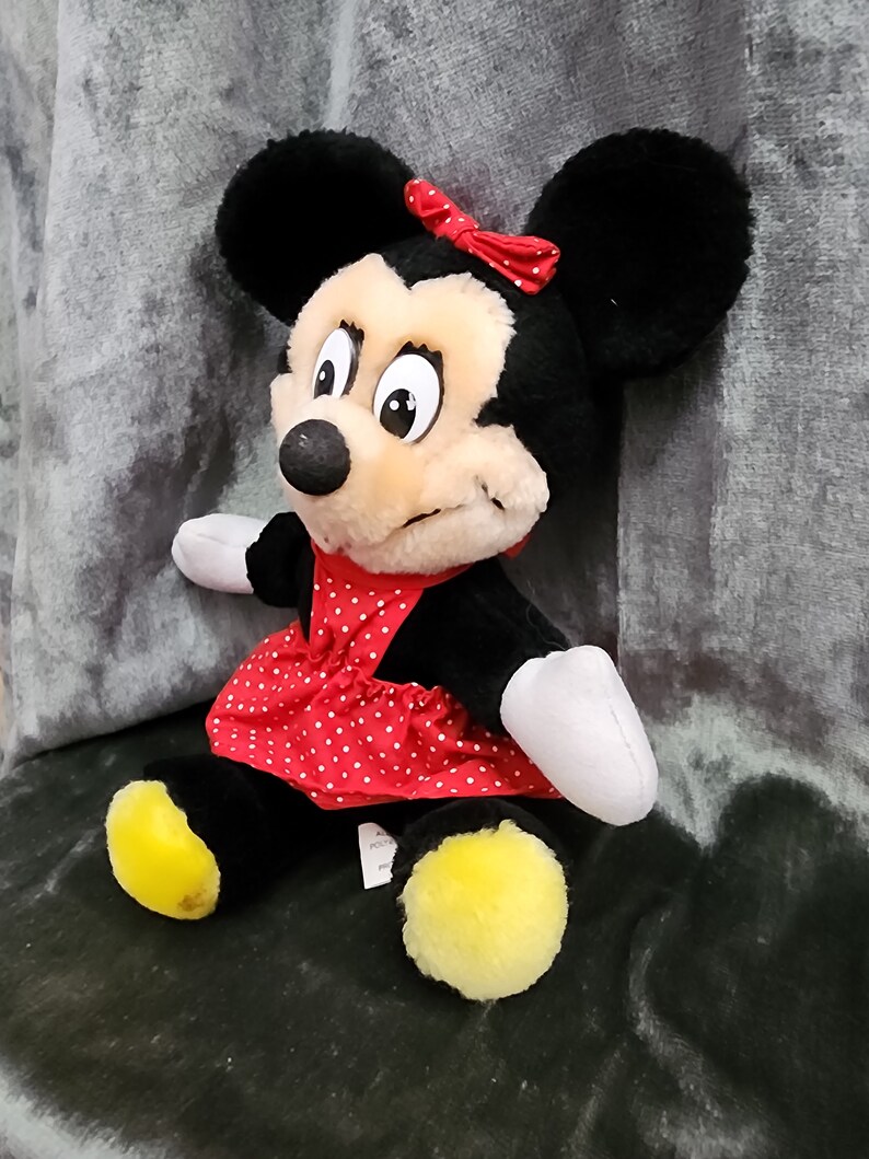 Puede incluir: Un juguete de peluche Minnie Mouse negro y tostado con un vestido rojo de lunares y un lazo rojo. El juguete tiene pies amarillos y est&aacute; sentado en una superficie gris.