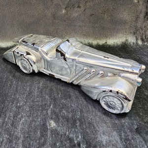 May include: A silver vintage toy car with a detailed design. The car has a long hood and a rounded body. The toy car is made of metal and has a worn finish.