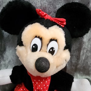 Puede incluir: Un juguete de peluche de Minnie Mouse con un vestido rojo con lunares blancos y un lazo rojo. Tiene pelo negro, ojos grandes y una nariz negra.