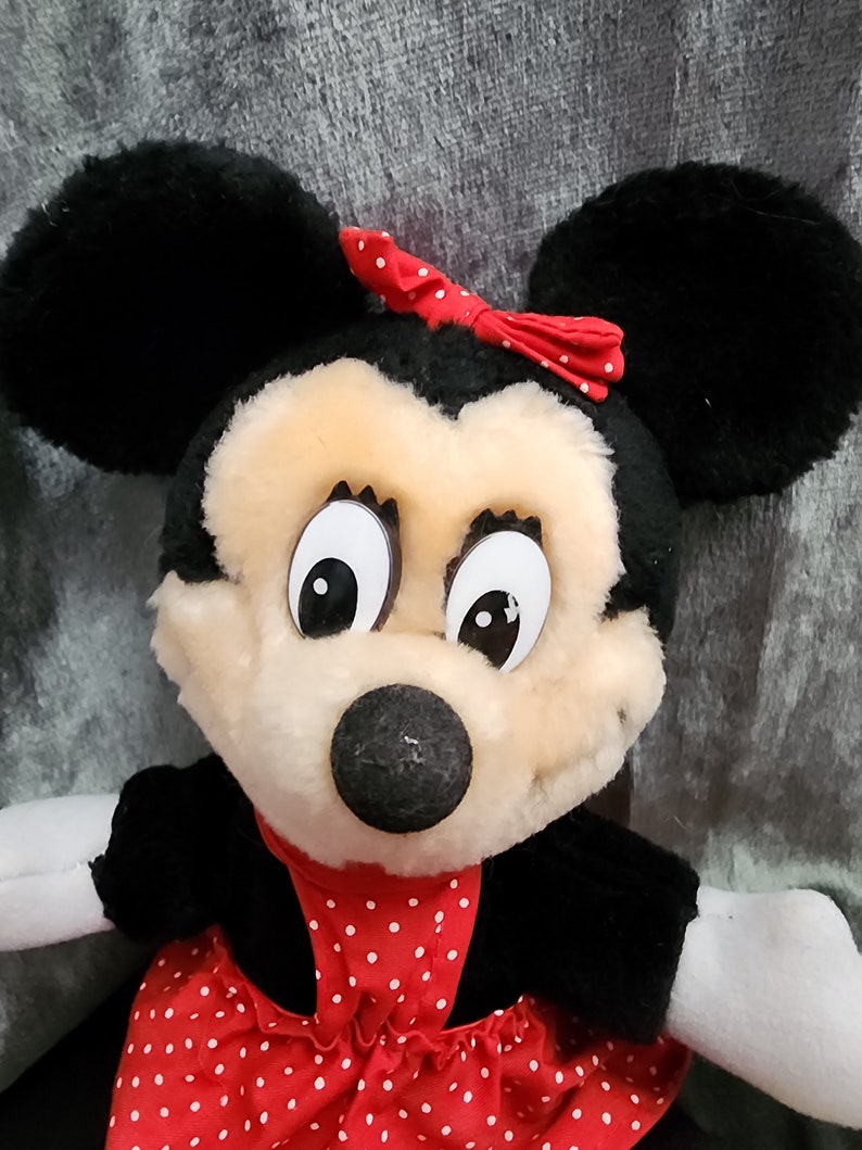 Puede incluir: Un juguete de peluche de Minnie Mouse con un vestido rojo con lunares blancos y un lazo rojo. Tiene pelo negro, ojos grandes y una nariz negra.