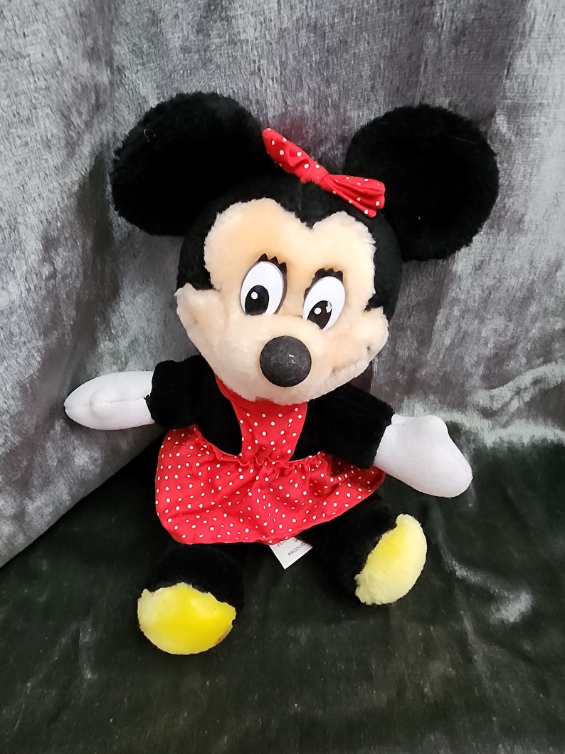Puede incluir: Un juguete de peluche de Minnie Mouse con un vestido rojo de lunares blancos y un lazo rojo. Tiene orejas negras y una nariz negra. Sus ojos son grandes y marrones.