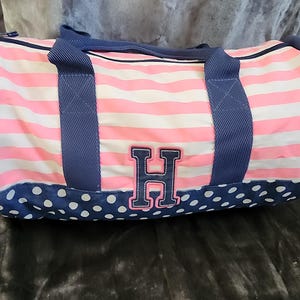 May include: Pink and white striped duffel bag with blue polka dots and a navy blue handle. The bag has a large navy blue letter "H" on the front.