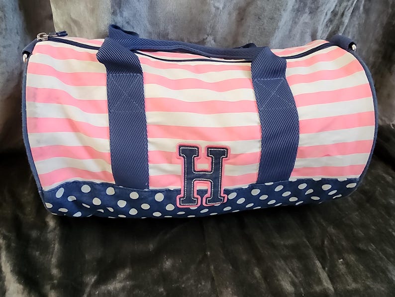 May include: Pink and white striped duffel bag with blue polka dots and a navy blue handle. The bag has a large navy blue letter "H" on the front.