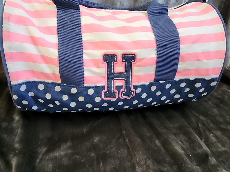 May include: A pink and white striped duffel bag with blue polka dots and a navy blue embroidered letter H on the front.