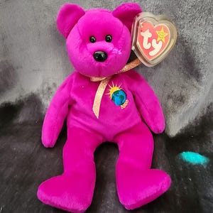 May include: A pink Ty Beanie Baby bear named "Princess" with a gold ribbon and a tag that says "Beanie Babies" and "Ty". The bear has a black nose and black eyes. The bear is wearing a gold ribbon with a small globe charm that says "2000".
