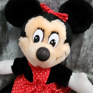 Puede incluir: Un juguete de peluche de Minnie Mouse con un vestido rojo con lunares blancos y un lazo rojo. Tiene orejas negras y una nariz negra.