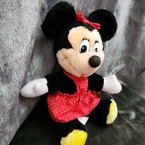 Puede incluir: Un juguete de peluche de Minnie Mouse con un vestido rojo con lunares blancos y un lazo rojo. Tiene orejas negras y una nariz negra.