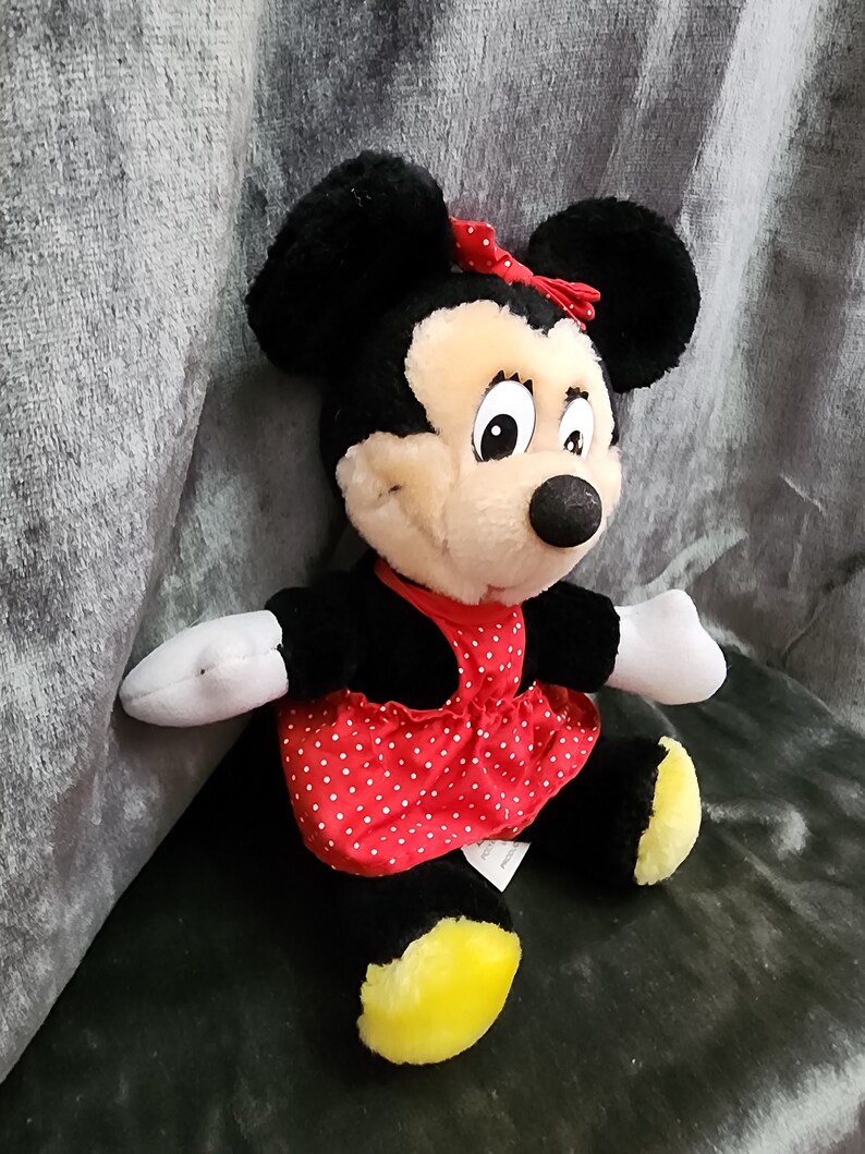 Puede incluir: Un juguete de peluche de Minnie Mouse con un vestido rojo con lunares blancos y un lazo rojo. Tiene orejas negras y una nariz negra.
