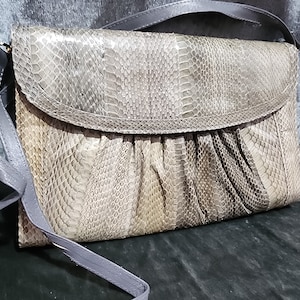 Puede incluir: Un bolso de mano de piel de serpiente gris con cierre de solapa y una correa larga. El bolso tiene una superficie texturizada y escamosa.