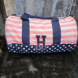 May include: Pink and white striped duffel bag with a blue polka dot bottom and a blue fabric handle. The bag has a large blue felt letter "H" on the front.