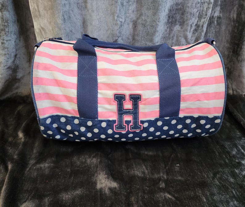 May include: Pink and white striped duffel bag with a blue polka dot bottom and a blue fabric handle. The bag has a large blue felt letter "H" on the front.