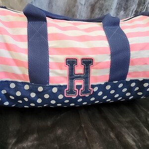 May include: A pink and white striped duffel bag with blue polka dots and a navy blue handle. The bag has a large navy blue and white letter "H" on the front.