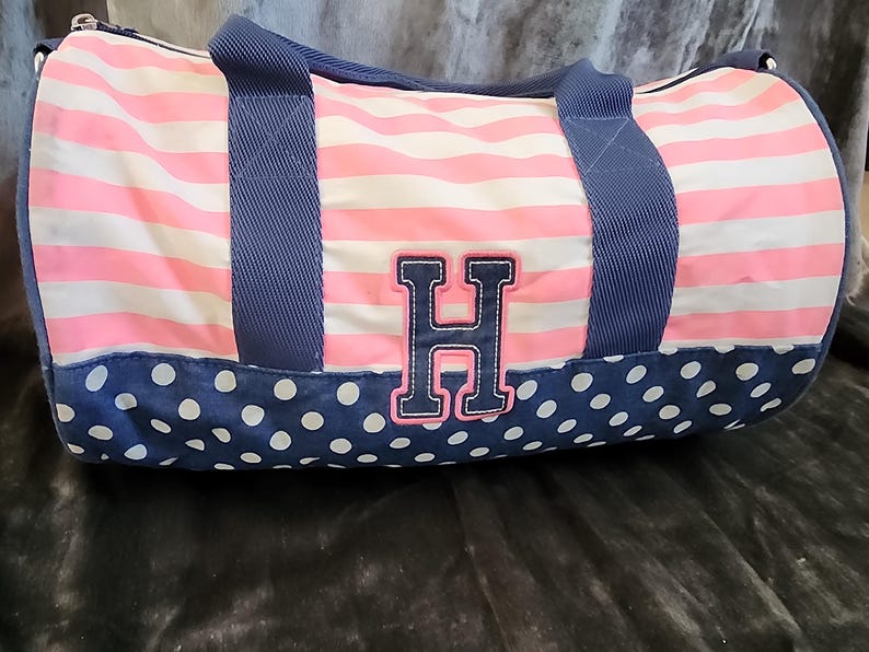 May include: A pink and white striped duffel bag with blue polka dots and a navy blue handle. The bag has a large navy blue and white letter "H" on the front.